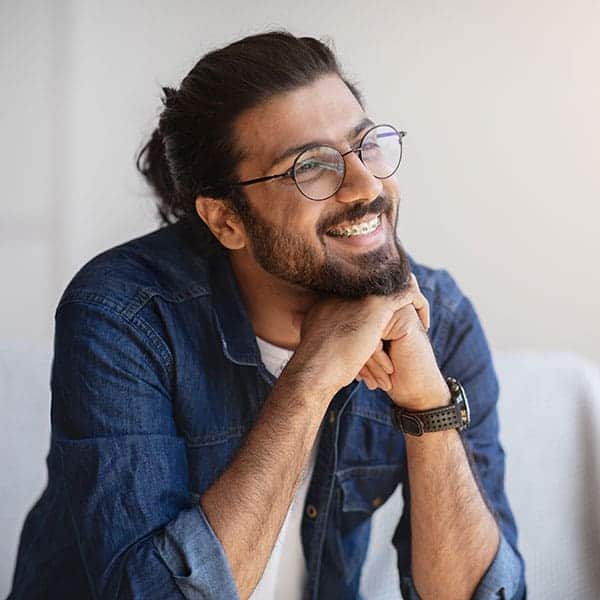 Smiling man with round glasses and a beard, wearing a denim shirt, resting his chin on his folded hands.