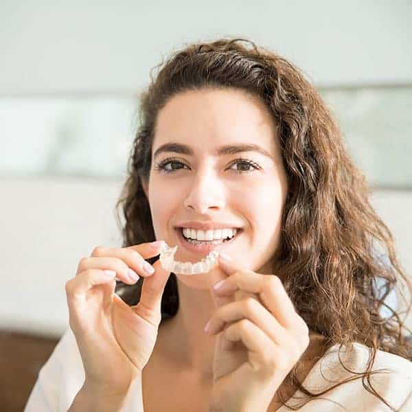 Smiling woman holds a clear dental aligner up to her teeth, showing it to the camera.