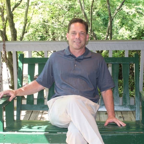 Smiling man in a gray polo sitting cross‑legged on a green wooden porch swing outdoors among trees and sunlight, facing the camera.
